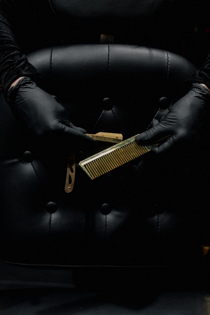 A barber with gloved hands holding a comb and razor against a leather chair backdrop.